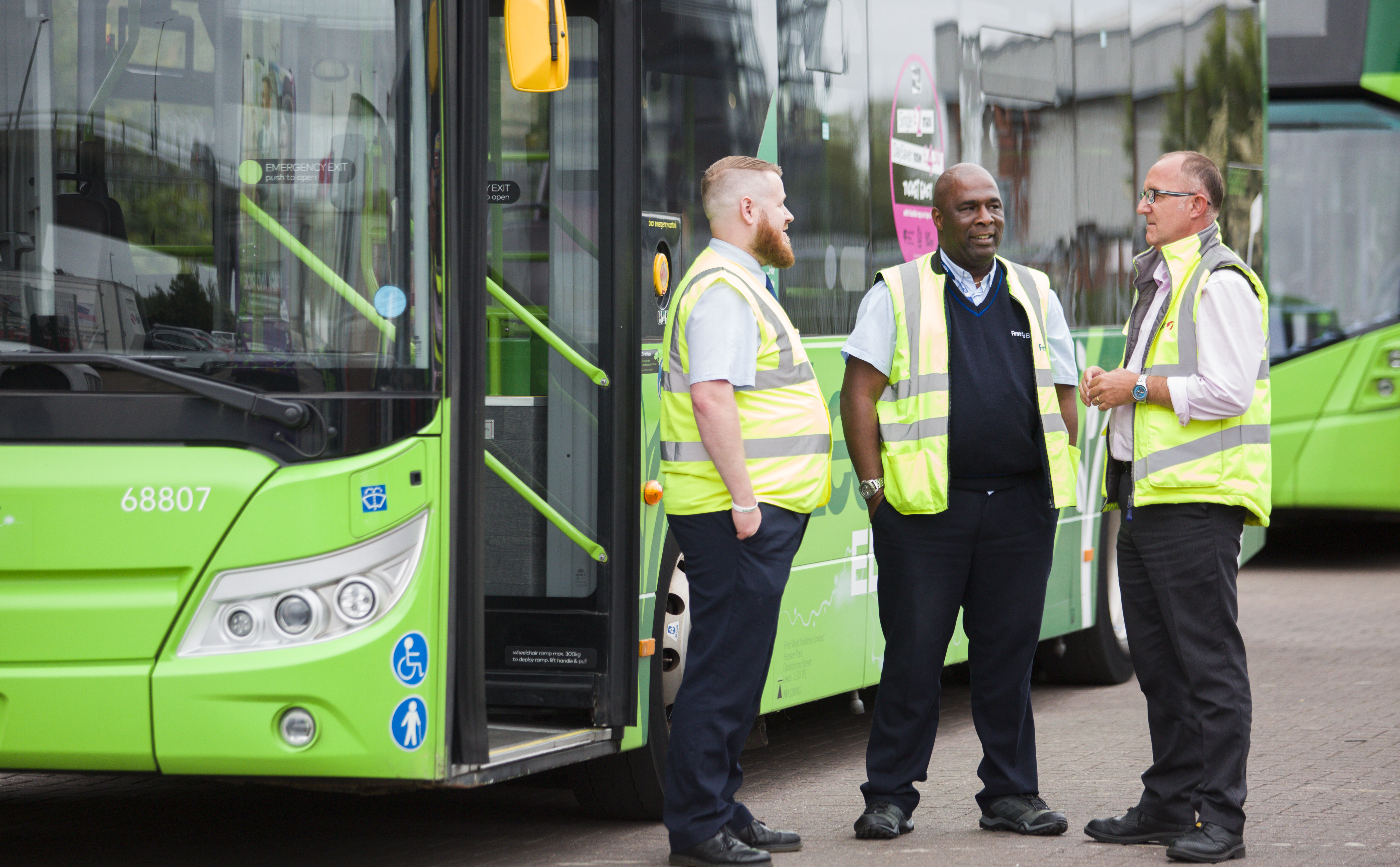 Three men in yellow high viz jackets stand talking in front of a green bus