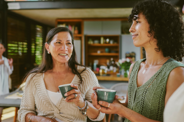 Several people gathered indoors in a modern kitchen setting, holding green ceramic cups. The background shows wooden shelves with various kitchen items, including bowls, jars, and plants.