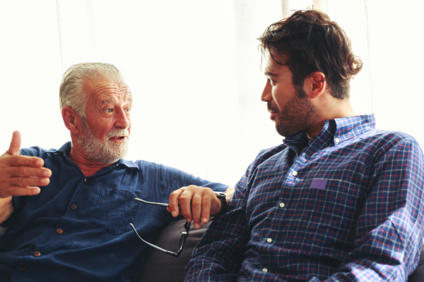 Two people sitting together on a sofa having a conversation. One gestures with his hand while holding a pair of glasses, and the other listens attentively. Soft natural light comes through a window behind them, creating a calm, informal setting.
