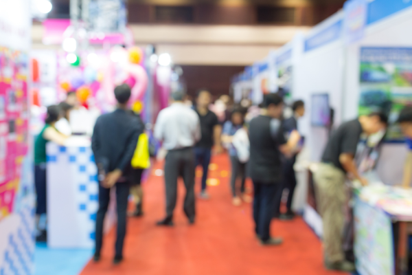 Blurred photo of an event with exhibiting stalls and individuals in conversation and walking around the event space
