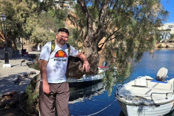 A man stood by a tree in Greece with a boat in the harbour